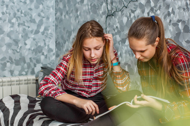 Two Teenage Girls reading fashion Magazine sitting on a couch at home on gray wall background - 写真・画像