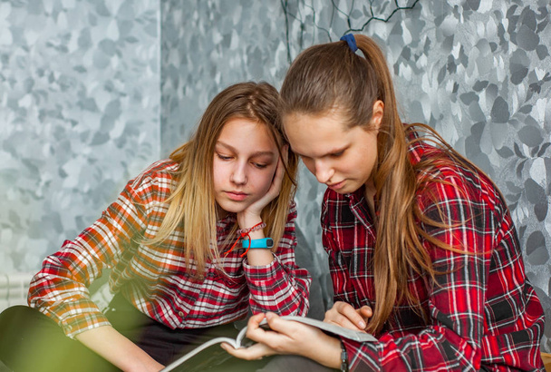 Two Teenage Girls reading fashion Magazine sitting on a couch at home on gray wall background - Fotoğraf, Görsel