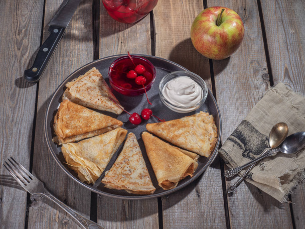 パンケーキは、大きな灰色のプレート上の三角形によってロールアップ, サワークリームと缶詰のサクランボと小さなアイスクリームボウルの隣に, 大きなリンゴ, チェリーと食器やナプキンの瓶が広がって  - 写真・画像
