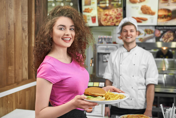 Woman keeping plate with fast food and smiling in cafe. - Foto, Imagem