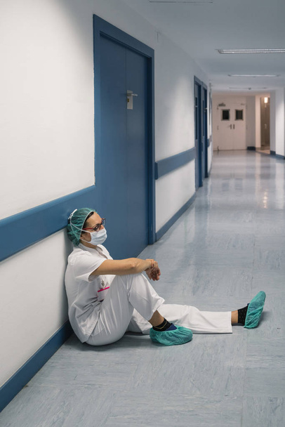 Vista lateral de la mujer en uniforme médico blanco apoyado en la pared mientras está sentado en el suelo en el hospital
 - Foto, imagen