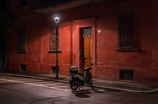 Noche en la ciudad. Scooter en el estacionamiento debajo de una farola cerca del edificio rojo
. - Foto, Imagen