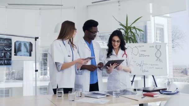 Medical team, two women and one african man standing and discuss diagnosis of the patient at hospital. Team of medical specialists discussing result of blood analysis patient in the conference room of - Кадры, видео