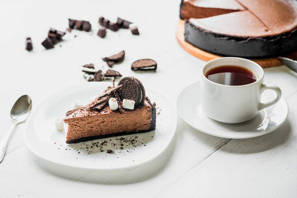 Chocolate cheesecake with pieces of chocolate, cookies and marshmallow on a white plate next to a cup of tea on the table - Foto, afbeelding