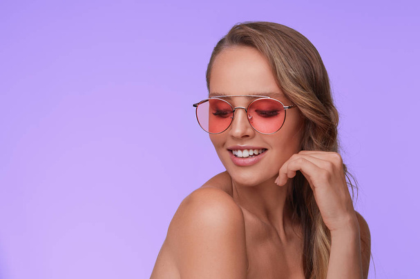 Portrait of beautiful young woman with perfect makeup wearing pink sunglasses. Smiling fashion model in aviator sunglasses posing on lilac background. Studio shot. Summer vacation. - Fotoğraf, Görsel