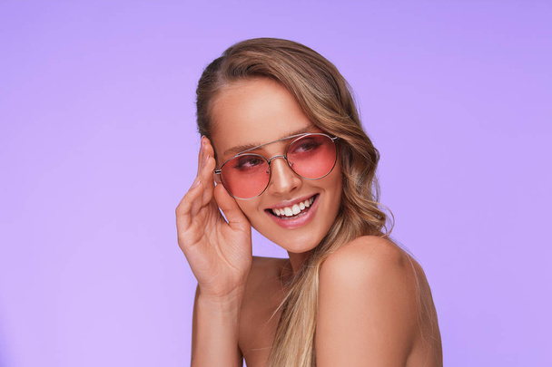 Portrait of beautiful young woman with perfect makeup wearing pink sunglasses. Smiling fashion model in aviator sunglasses posing on lilac background. Studio shot. Summer vacation. - Photo, image