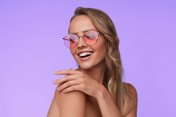 Portrait of beautiful young woman with perfect makeup wearing pink sunglasses. Smiling fashion model in aviator sunglasses posing on lilac background. Studio shot. Summer vacation. - Foto, Imagem