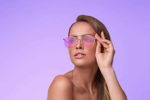 Portrait of beautiful young woman with perfect makeup wearing pink sunglasses. Smiling fashion model in aviator sunglasses posing on lilac background. Studio shot. Summer vacation. - 写真・画像