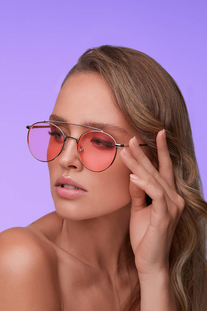 Portrait of beautiful young woman with perfect makeup wearing pink sunglasses. Smiling fashion model in aviator sunglasses posing on lilac background. Studio shot. Summer vacation. - Фото, изображение