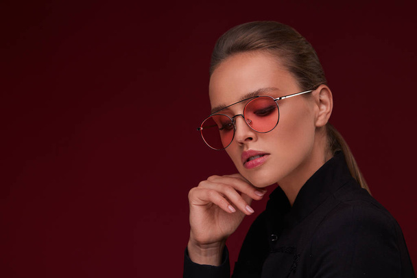 Portrait of beautiful young woman with perfect makeup wearing pink sunglasses. Smiling fashion model in aviator sunglasses posing on burgundy background. Studio shot. Summer vacation. - Valokuva, kuva