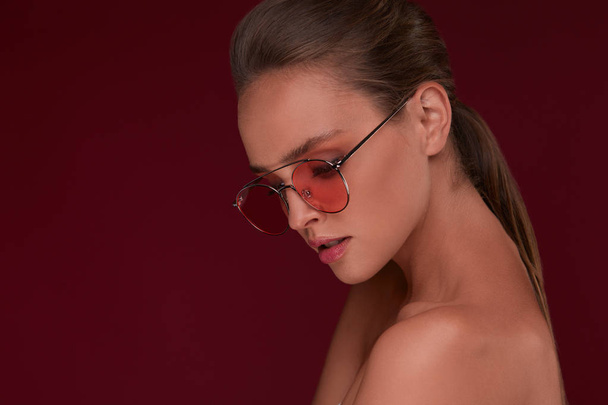 Portrait of beautiful young woman with perfect makeup wearing pink sunglasses. Smiling fashion model in aviator sunglasses posing on burgundy background. Studio shot. Summer vacation. - Foto, afbeelding