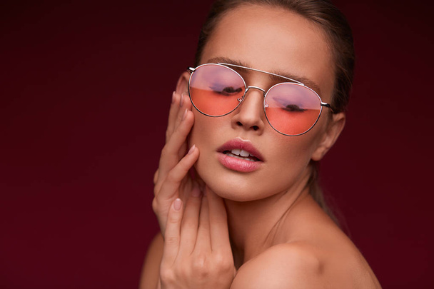 Portrait of beautiful young woman with perfect makeup wearing pink sunglasses. Smiling fashion model in aviator sunglasses posing on burgundy background. Studio shot. Summer vacation. - Foto, afbeelding