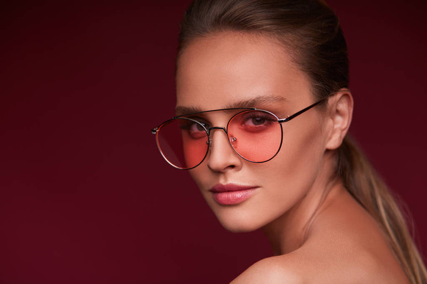 Portrait of beautiful young woman with perfect makeup wearing pink sunglasses. Smiling fashion model in aviator sunglasses posing on burgundy background. Studio shot. Summer vacation. - Photo, Image