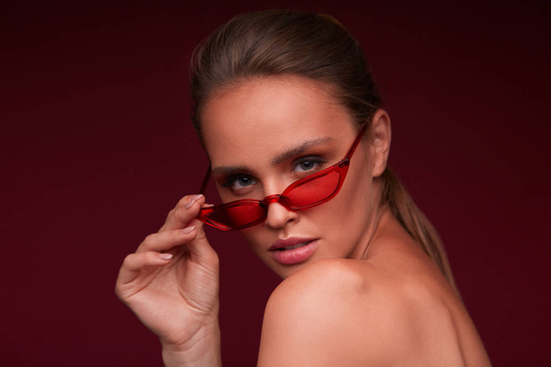 Portrait of beautiful young woman with perfect makeup wearing red sunglasses. Smiling fashion model in cat eye sunglasses posing on burgundy background. Studio shot. Summer vacation. - Φωτογραφία, εικόνα