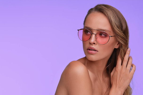 Portrait of beautiful young woman with perfect makeup wearing pink sunglasses. Smiling fashion model in aviator sunglasses posing on lilac background. Studio shot. Summer vacation. - Φωτογραφία, εικόνα