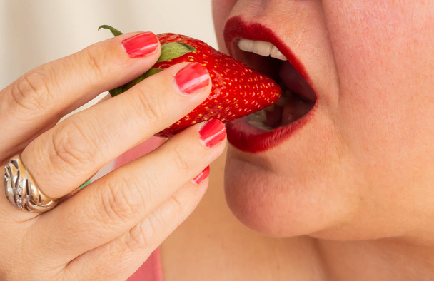 Een volwassen vrouw met rode nagels en lippen het eten van een aardbei - Foto, afbeelding