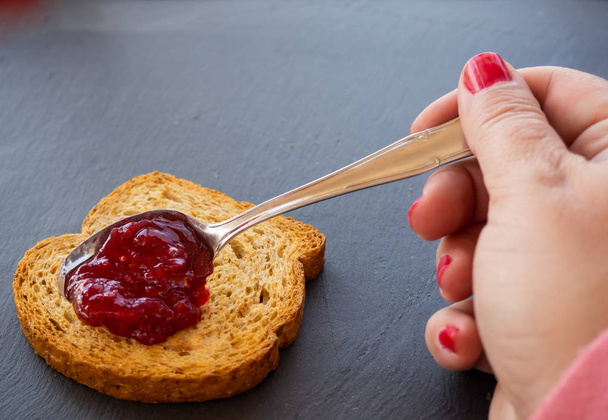 Une femme aux ongles peints enduisant de confiture de framboise rouge avec une cuillère sur une tranche de pain grillé
 - Photo, image