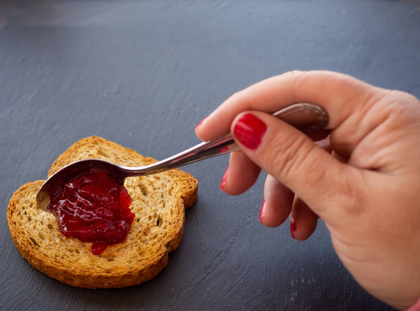 トーストのスライスにスプーンで赤いラズベリージャムを塗った爪を持つ女性 - 写真・画像