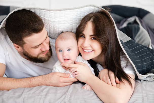 Jovem família sorridente com bebê sob cobertor
 - Foto, Imagem