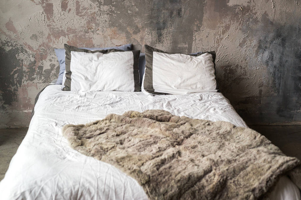 Bed with pillows and fur blanket against grungy plaster wall - Photo, image