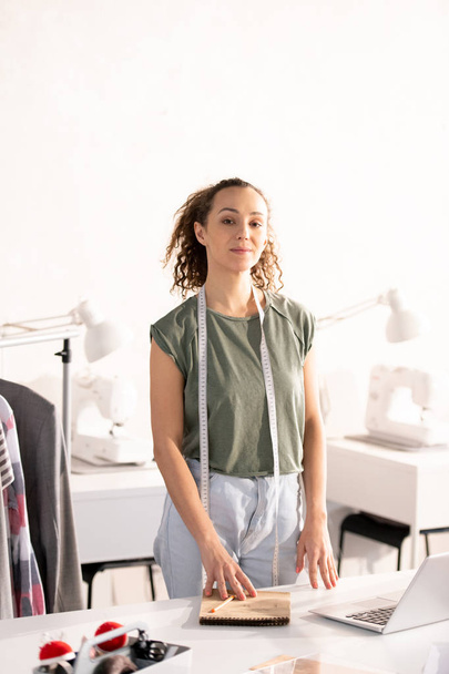 junge lässige Designerin einer Modekollektion am Schreibtisch vor der Kamera in ihrer Werkstatt oder im Atelier - Foto, Bild