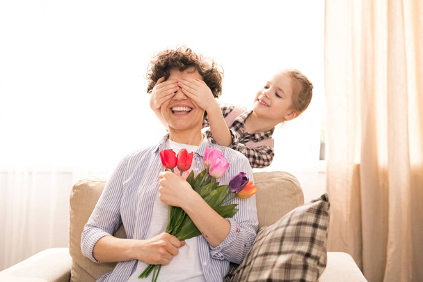 Rindo jovem mãe com um monte de tulipas sentadas em poltrona e adivinhando quem cobrindo os olhos com as mãos
 - Foto, Imagem