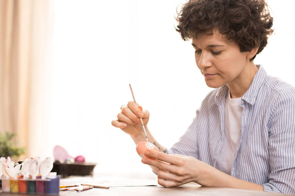 Young brunette woman with short curly hair holding egg and paintbrush while painting Easter ornament - Foto, imagen