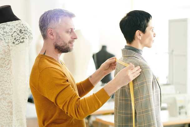 Sastre profesional maduro tomando medidas de chaqueta mientras trabaja con su cliente en sastrería
 - Foto, Imagen
