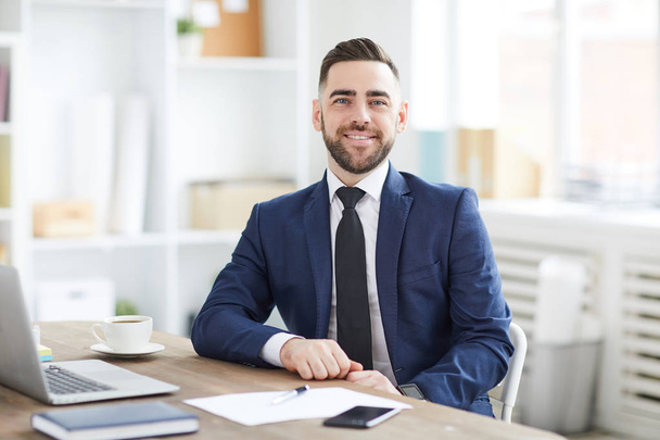 Joven hombre de negocios alegre atractivo en traje y corbata mirándote desde su lugar de trabajo en la oficina
 - Foto, imagen