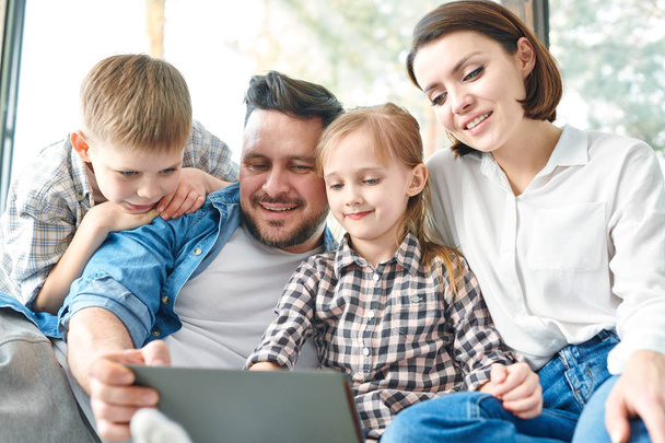 Szczęśliwa młoda Współczesna rodzina czterech patrząc na wyświetlaczu Touchpad podczas oglądania ciekawych rzeczy w sieci - Zdjęcie, obraz