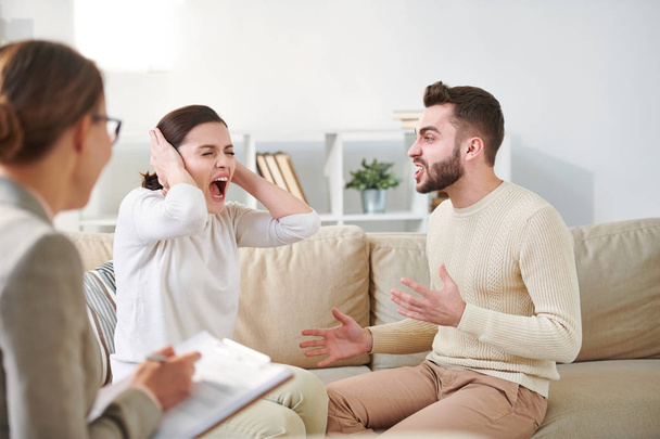 Giovane donna infastidita urlando e coprendo le orecchie per mano mentre il marito nervoso cerca di spiegare qualcosa durante la sessione di famiglia
 - Foto, immagini