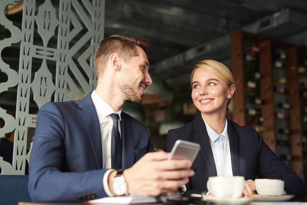 Twee jonge eigentijdse collega's in formalwear bespreken iets in de smartphone van zakenman in café - Foto, afbeelding