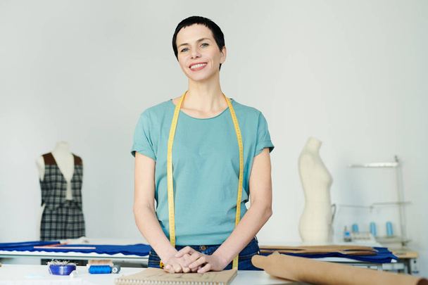 Σύγχρονη επαγγελματική γυναικεία σχεδιάστρια μόδας με ταινία μέτρησης στέκεται από το χώρο εργασίας μπροστά από την κάμερα - Φωτογραφία, εικόνα
