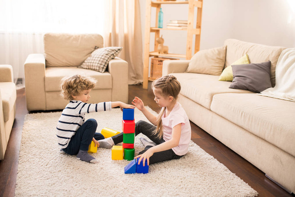 Fratelli svegli che giocano cubi di plastica di vari colori mentre siedono sul pavimento del soggiorno o all'asilo
 - Foto, immagini