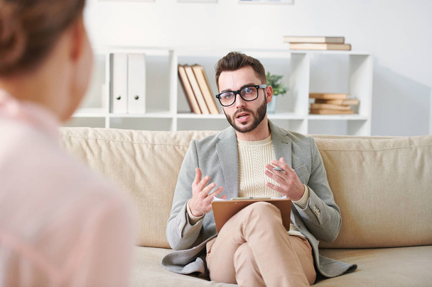 Confident counselor in smart casual and eyeglasses sitting on couch in front of patient and explaining his viewpoint about the situation - Photo, Image