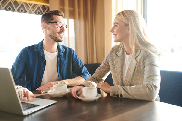 Gelukkige jonge elegante blonde zakenvrouw en haar collega in casualwear discussiëren over werk momenten met kopje thee in café - Foto, afbeelding