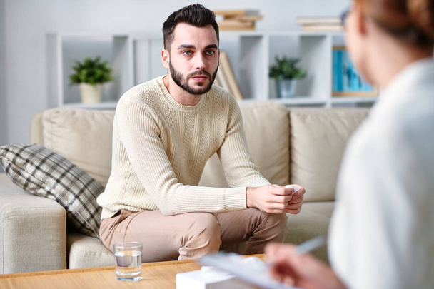 Серьезный молодой пациент психотерапевта, сидящий на диване и слушающий ее советы во время обсуждения его проблем на работе
 - Фото, изображение