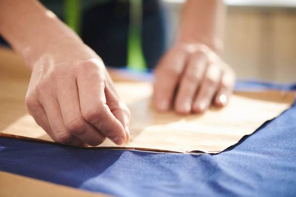 Το χέρι της νεαρής ράφτρα καρφιτσώνει χαρτί σε μπλε ύφασμα πριν το περικυκλώσει - Φωτογραφία, εικόνα