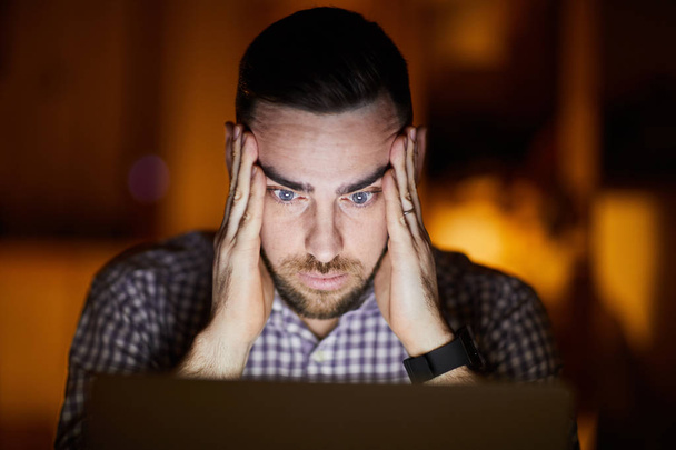 Tense or shocked businessman in casualwear touching his temples while looking at laptop display in darkness - 写真・画像