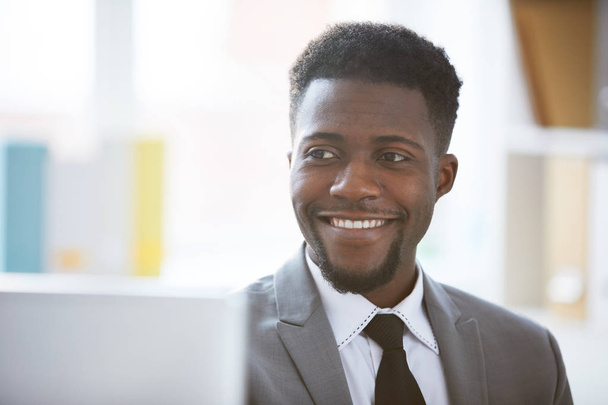 Счастливый молодой бизнесмен с зубастой улыбкой сидит перед ноутбуком или монитором компьютера в офисе
 - Фото, изображение