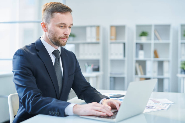 Молодий бородатий бізнесмен у формальному одязі сидить за столом в офісі перед ноутбуком в робочий день
 - Фото, зображення