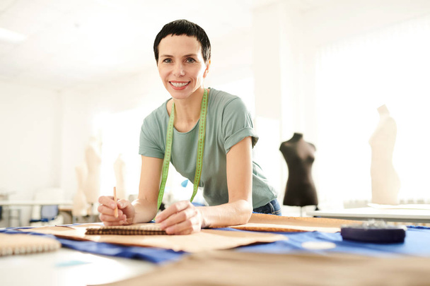 glückliche brünette Schneiderin in Freizeitkleidung beugt sich über ihren Arbeitsplatz, während sie Skizzen und andere Arbeiten im Modestudio macht - Foto, Bild