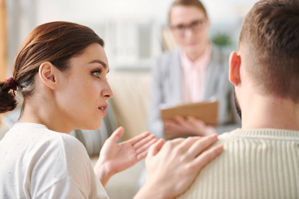 Молодая женщина смотрит на своего мужа, объясняя что-то перед своим консультантом во время семейной консультации
 - Фото, изображение