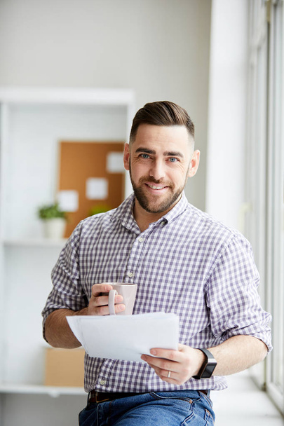 Молодий успішний бізнесмен в повсякденному одязі п'є під час перегляду фінансового документа в офісі
 - Фото, зображення