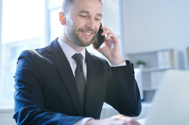 Улыбающийся молодой бизнесмен в элегантном костюме смотрит на вывеску lapto pdisplay во время общения и общения со смартфоном
 - Фото, изображение
