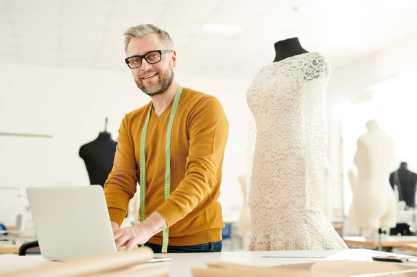 Érett sikeres Tailor a casualwear állva az asztalra, és keresi az új kreatív ötletek és divatirányzatok a neten - Fotó, kép