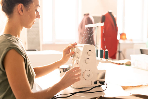 Nuori nainen satunnaisissa vaatteissa istuu sähköompelukoneen vieressä ja valmistelee sitä työhön
 - Valokuva, kuva
