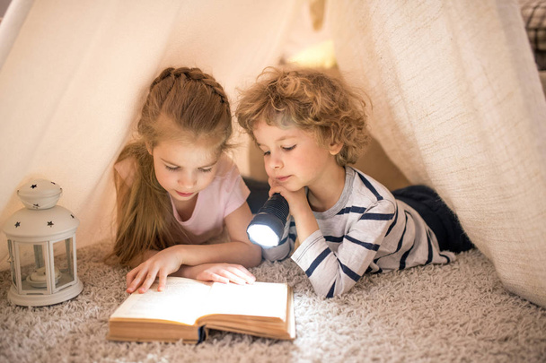 グループの2つのかわい子が床に自作テントの中に横たわって、物語の本を読んでいる間にランタンを使用する - 写真・画像