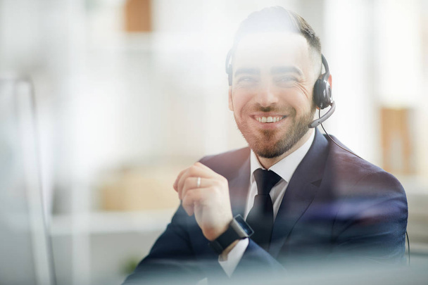 Cheerful young elegant operator in headset talking to clients and consulting them while sitting in office - Фото, зображення