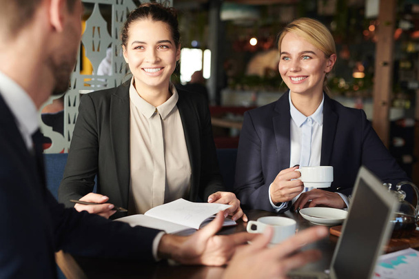 Dwóch szczęśliwych udanych kobiet pracowników w formalwear słuchania ich kolega wyjaśnia strategię biznesową na spotkaniu w kawiarni - Zdjęcie, obraz